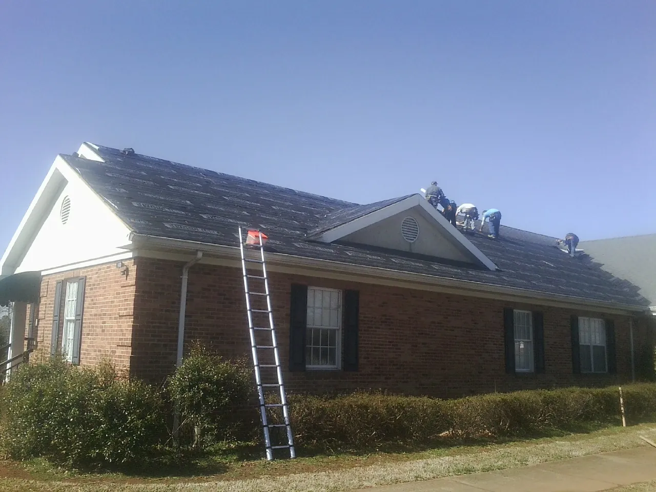 Crew tearing shingles off a brick ranch roof with a ladder set — Emergency Roof Repair work by our crew in Earlham