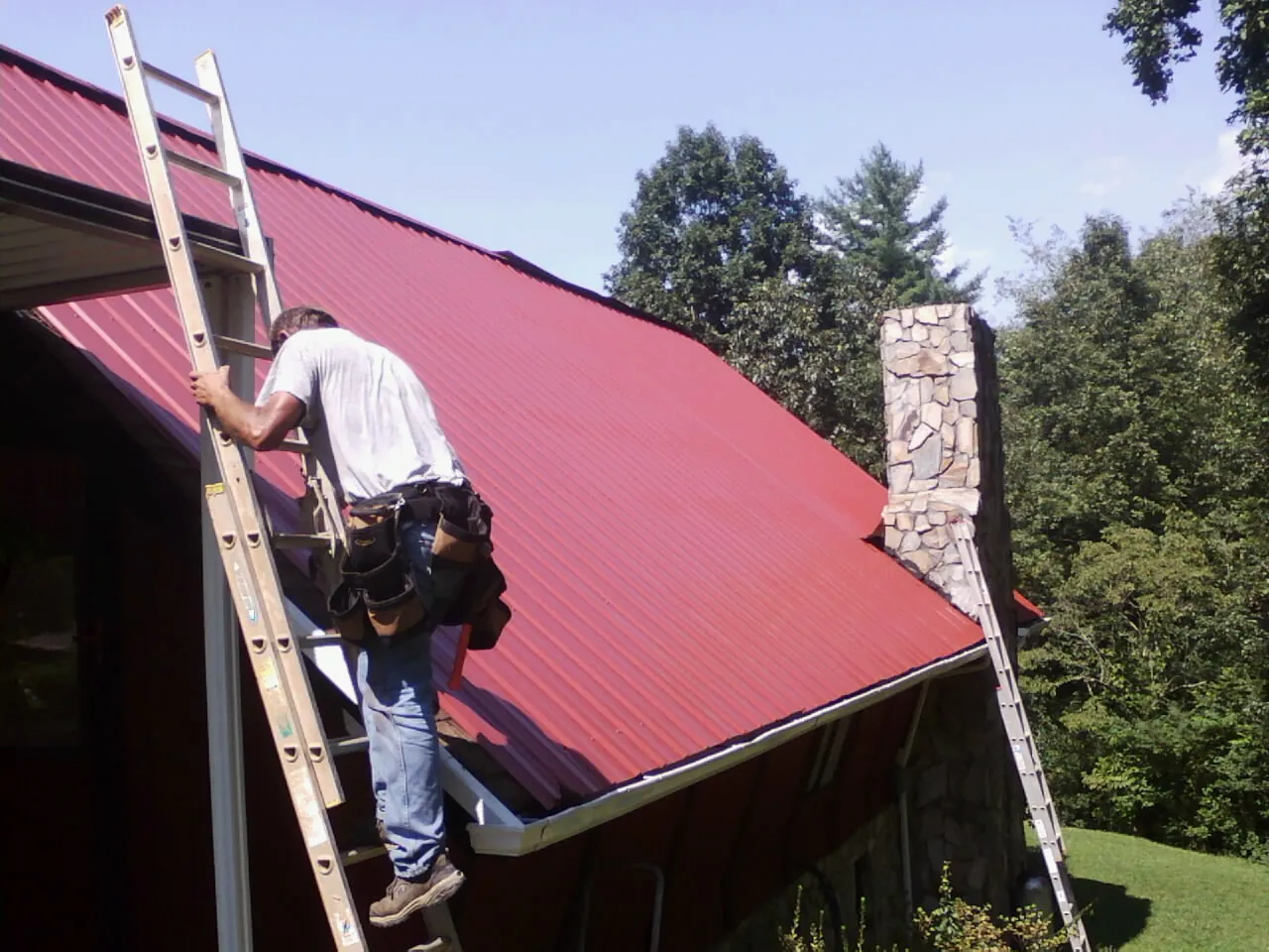 Roofer on a ladder beside a red metal roof and stone chimney — Soffit & Fascia Repair by Roofing Brothers in Earlham